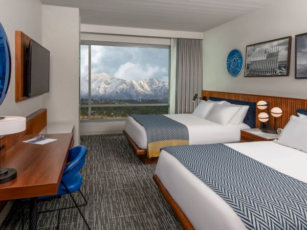 A modern hotel room with two beds, a desk, TV, large window showing snow-capped mountains, and blue and white decor, ends the sentence.