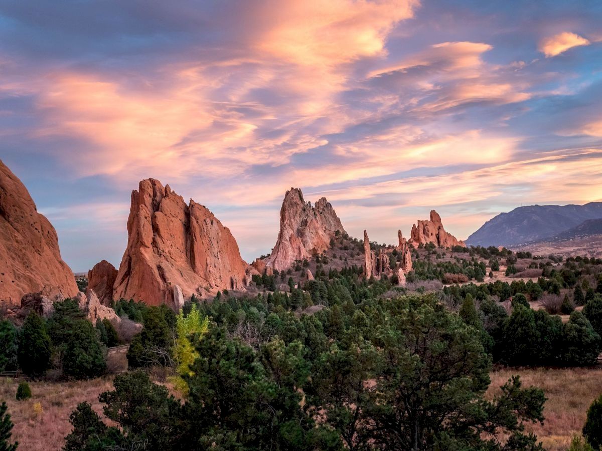 Desert landscape with rugged pink rock formations, green shrubs, and a dramatic sunset sky casting warm colors across the scene.