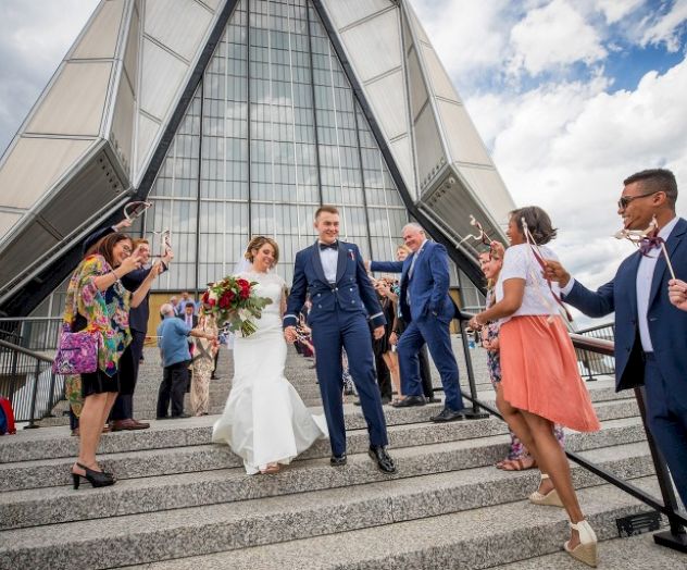 A newlywed couple walks down steps, surrounded by cheering friends and family, outside a modern architectural building under a partly cloudy sky.