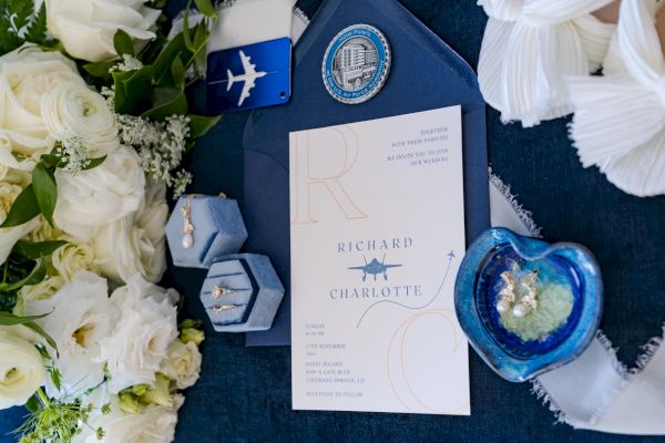 Wedding invitation with floral arrangement, jewelry boxes, and decorative items. The invitation features names and an airplane motif.