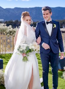 A bride and groom walk outside, with mountains in the background. The bride holds a bouquet and wears a veil.