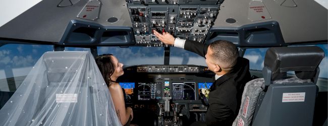 A bride and groom in a flight simulator, with the bride wearing a veil and the groom reaching for controls, surrounded by cockpit panels.