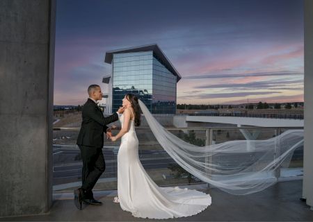 A couple in wedding attire stands on a balcony with the cityscape and a modern building in the background, during a colorful sunset.