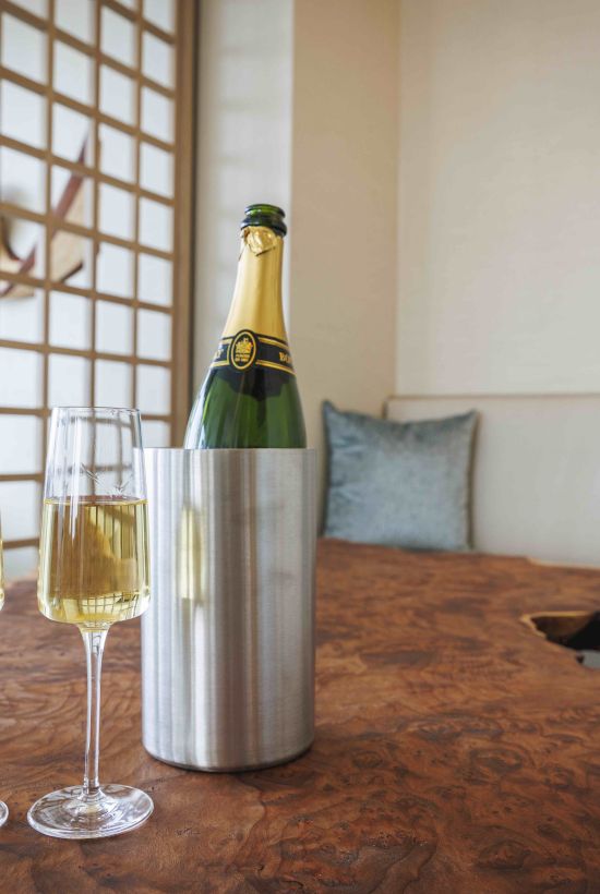 Champagne bottle in an insulated sleeve with two flutes on a rustic table, a cozy white lounge and decorative screen in the background.