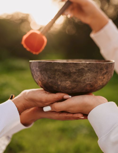 Two people hold a bowl while one sprinkles with a small brush; looks like a ceremonial ritual or offering in a sunlit outdoor setting.