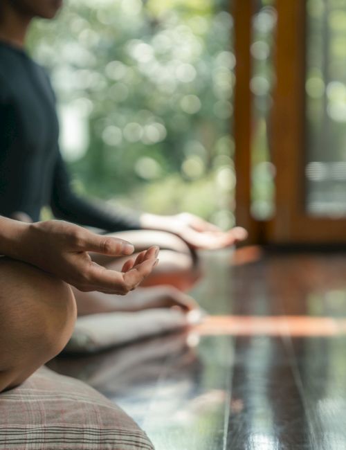 People meditating indoors on a wooden floor near an open door, seated cross-legged with hands resting on knees.