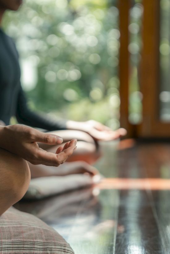 People meditating indoors on a wooden floor, sitting cross-legged with hands resting on knees, soft natural light from an open door.