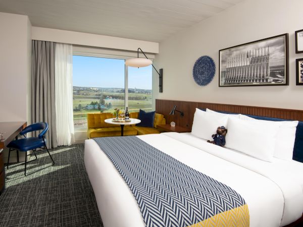 A modern hotel room with a double bed, a blue chair, yellow sofa, and a window view. The decor includes framed pictures and blue accents.
