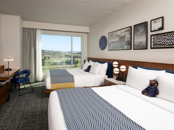 A hotel room with two beds, wall art, a desk with a chair, and a view through a large window.