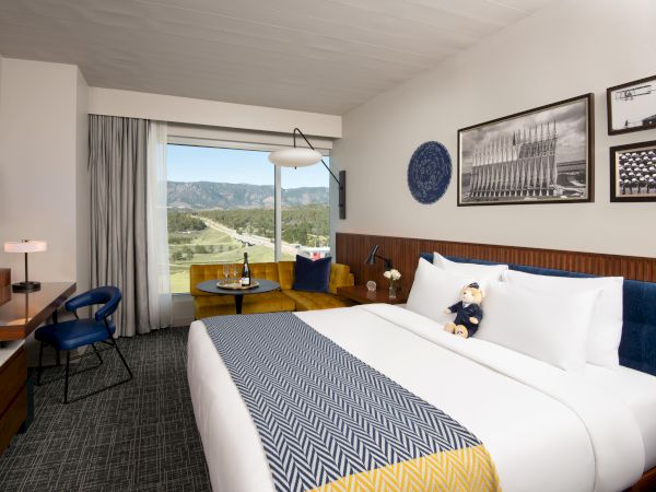 A hotel room features a bed with a patterned blanket, a sofa, a table with wine, art on walls, and a large window offering scenic views.