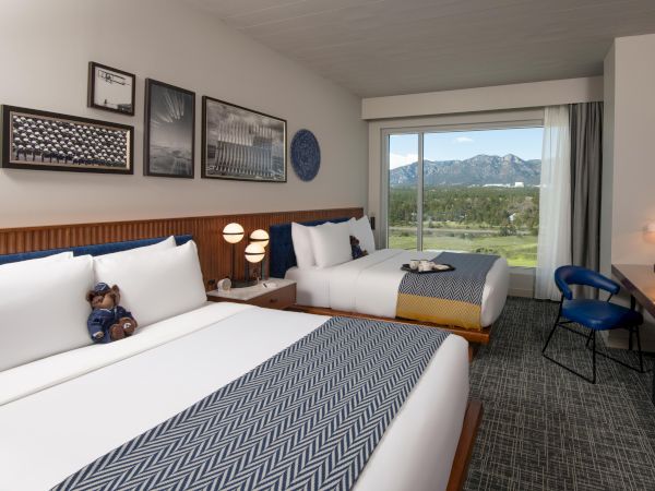This image shows a hotel room with two beds, a window with a scenic view, artwork on the wall, and a desk with a chair.
