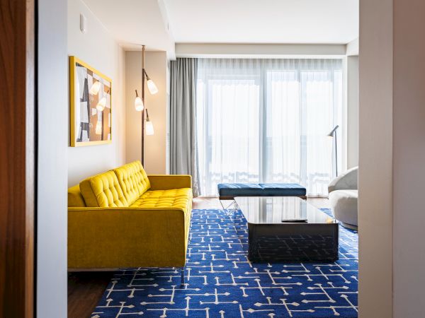 A modern living room features a yellow sofa, geometric rug, and large windows with sheer curtains, creating a bright atmosphere.
