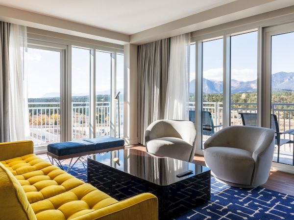 A modern living room with a yellow couch, two gray chairs, a black coffee table, and large windows showing a mountain view.