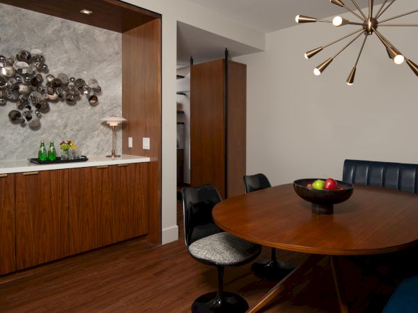 A stylish dining area features a modern light fixture, wooden table, black chairs, decorative wall art, and a sideboard with drinks.