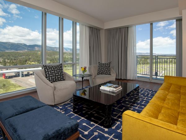 A modern living room with a yellow sofa, gray chairs, black coffee table, and large windows showing a scenic mountain view.