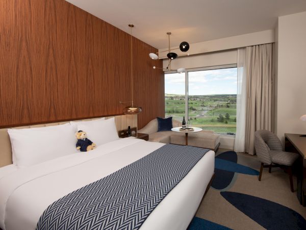 A cozy hotel room with a bed, teddy bear, desk, chair, stylish lamp, and window view of greenery outside completes the scene.