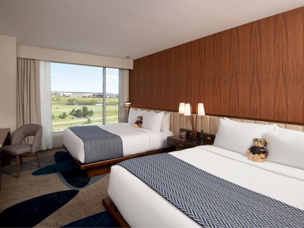 Two beds with teddy bears in a hotel room with a large window and a scenic view outside.
