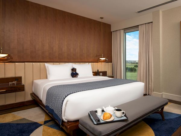 A cozy hotel room with a large bed, a teddy bear, a tray with breakfast, window view, and modern decor.