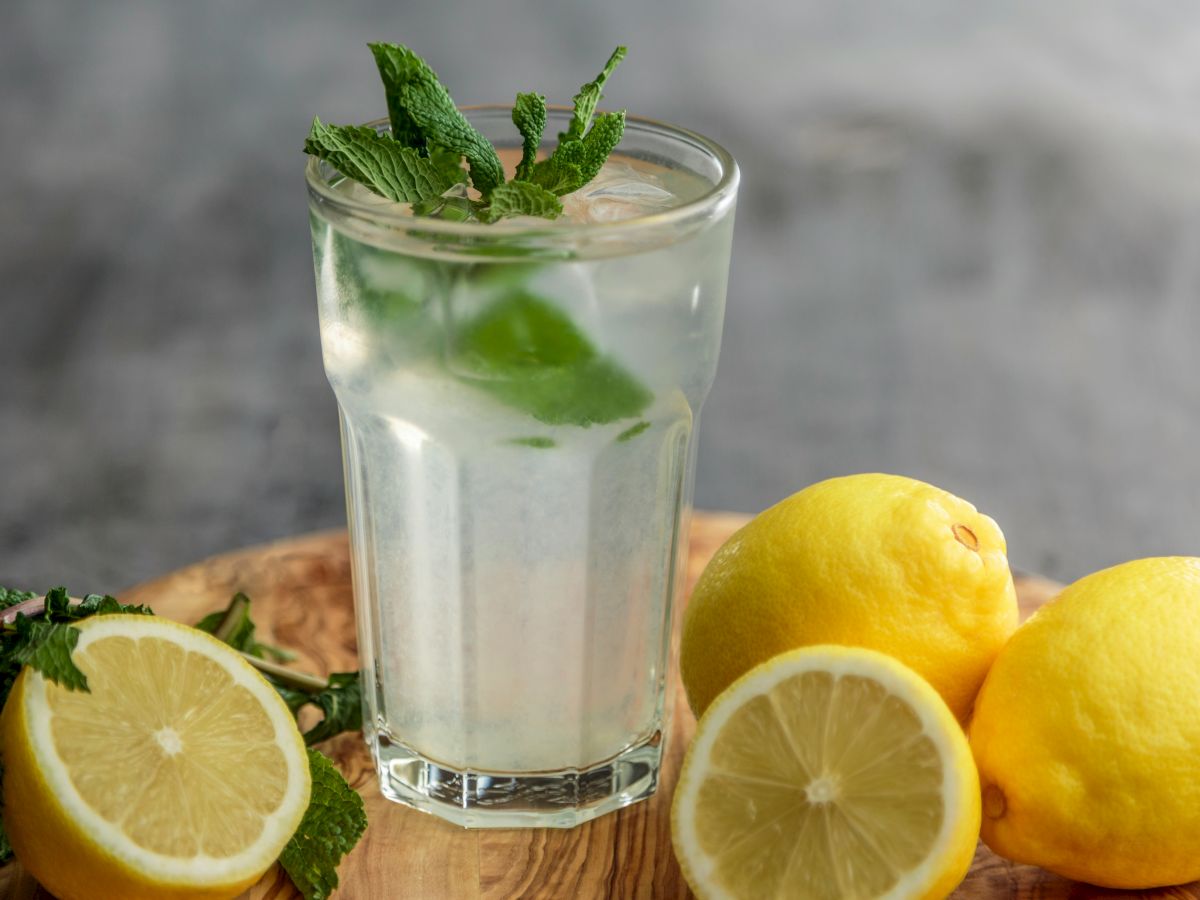 A glass of refreshing lemonade garnished with mint leaves, alongside fresh whole and sliced lemons and extra mint on a wooden board.