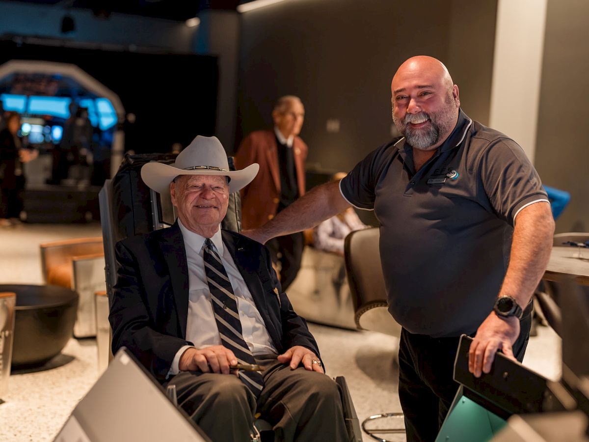 Two men in a showroom or tech space: one seated in a chair wearing a suit and cowboy hat, the other standing with a laptop, smiling; modern equipment and screens around.