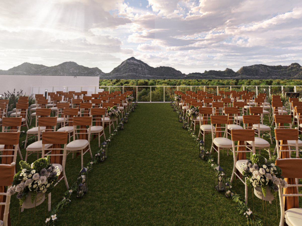 The image shows an outdoor wedding setup with rows of chairs on a lawn, mountains in the background, and a tree to the right.