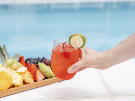 A hand reaches for a citrusy pink drink beside a platter of fresh fruit by a bright poolside&mdash;summery and refreshing at the edge of the water.