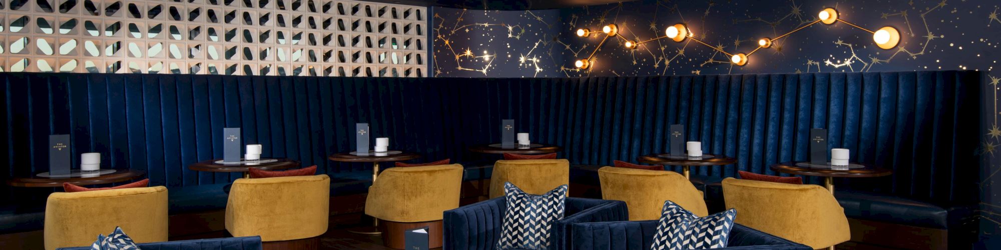 The image shows a stylish, modern lounge with plush blue seating, yellow chairs, patterned cushions, and ambient lighting.