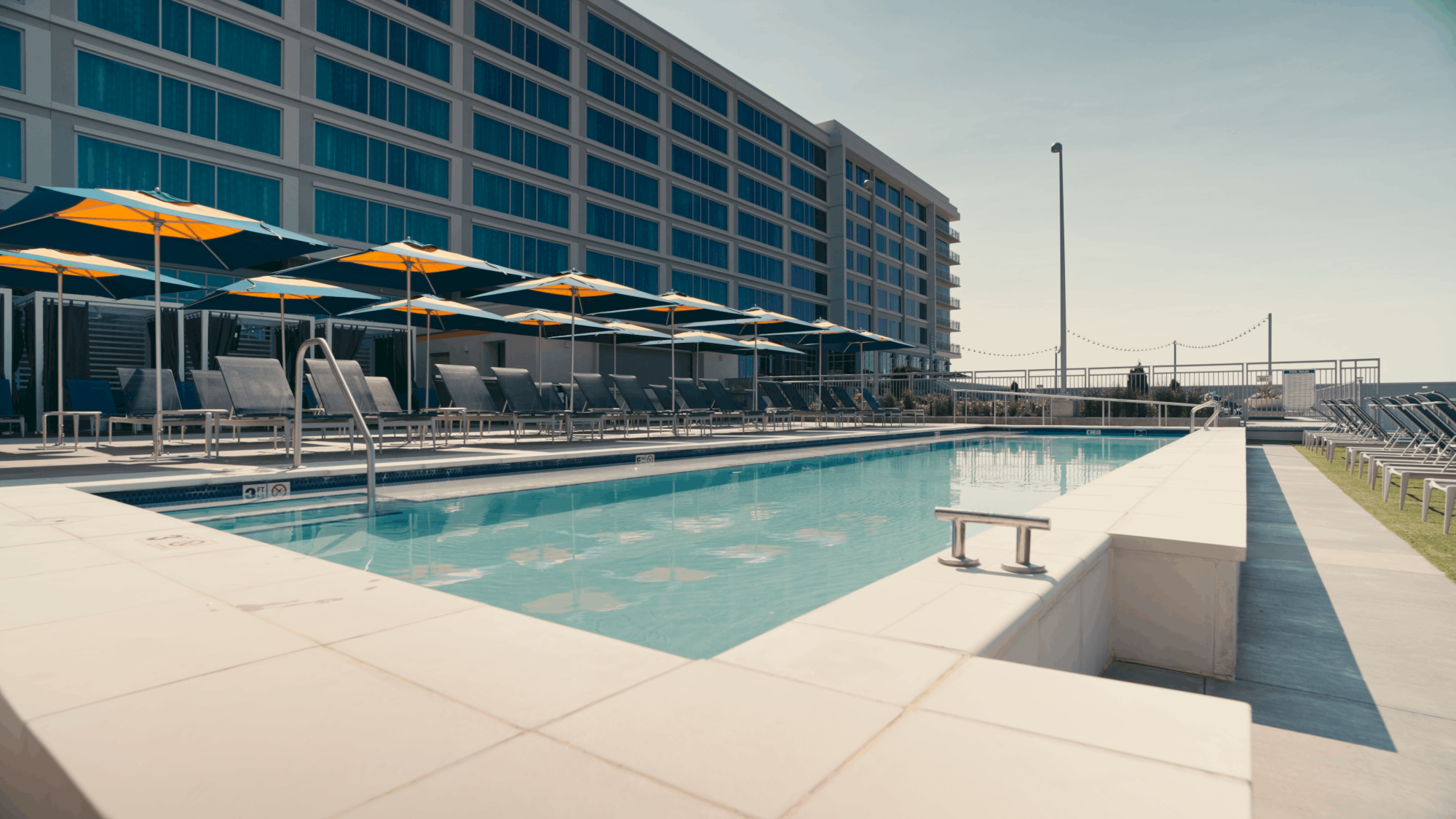 A modern hotel pool area with lounge chairs, yellow umbrellas, and a long rectangular pool beside a multi-story building, sunny and inviting.