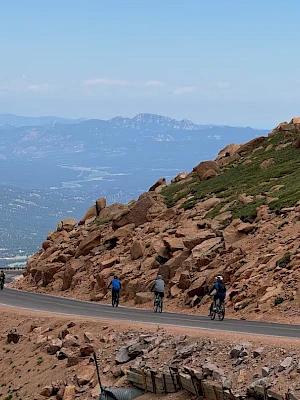 A winding mountain road with rocky slopes and a few cyclists pedaling along, distant valley and blue sky in the background.