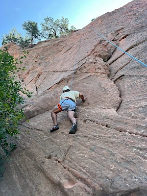 A climber wearing a helmet and harness ascends a steep rock face with a rope, gripping rough grooves and cracks in a sunlit cliff.