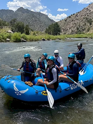 A group of six people wearing life jackets in a blue raft paddling down a rocky river surrounded by green hills and mountains.