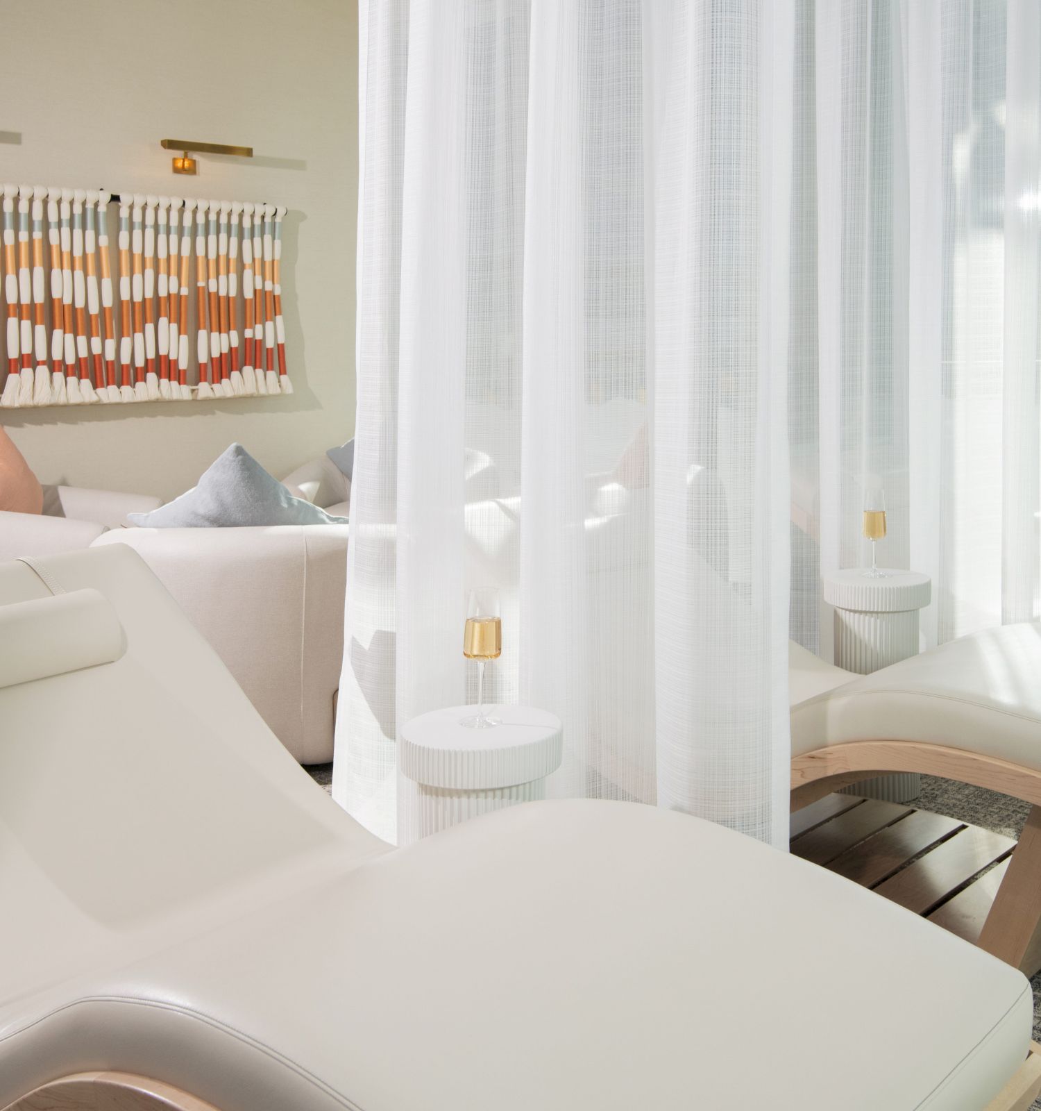 A serene spa lounge with white chaise lounges, sheer curtains, and soft natural light by a window, creating a tranquil retreat.