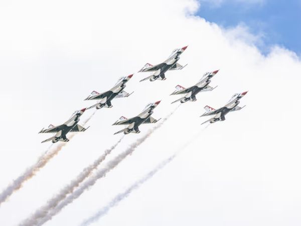 Six fighter jets fly in a tight formation, leaving colorful smoke trails in their wake as they ascend through a partly cloudy sky.