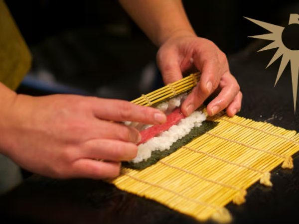 A person rolls sushi on a bamboo mat, placing filling along nori with white rice; a stylized sun logo sits in the corner.