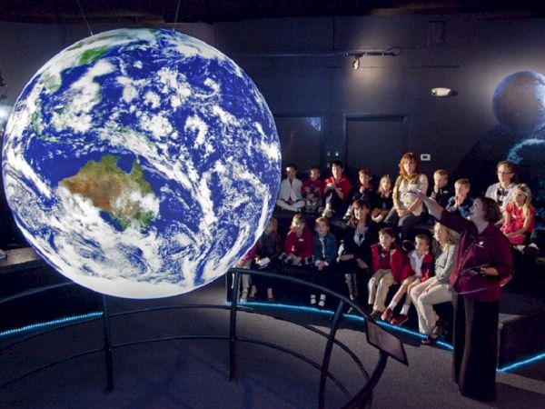 A group of people seated in a dark room watch a large, floating globe of Earth, with a second Moon-like globe in the background.