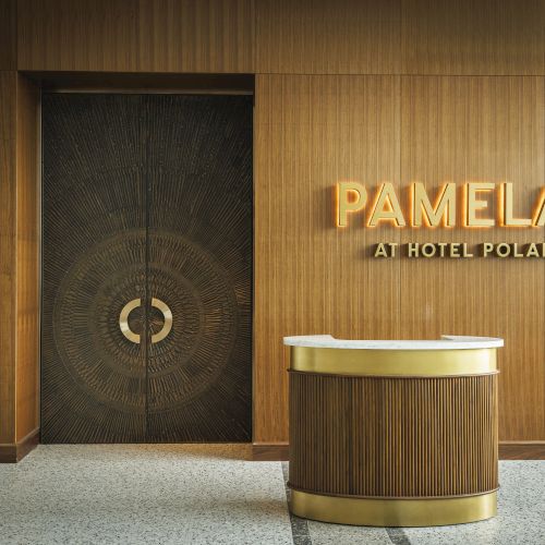 A wooden reception desk is in front of a wall with the illuminated sign "PAMELA'S AT HOTEL POLARIS" next to a decorative door.