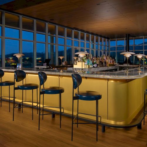 The image shows a stylish bar with a long marble counter, surrounded by high chairs, featuring dim lighting and bottles neatly arranged.