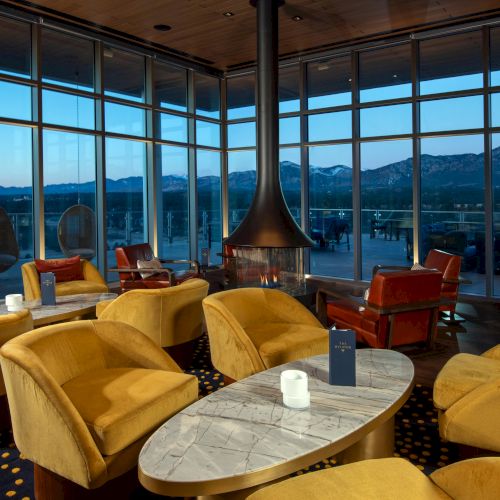 A cozy lounge with mustard-yellow chairs, a central fireplace, and large windows showcasing a mountain view.