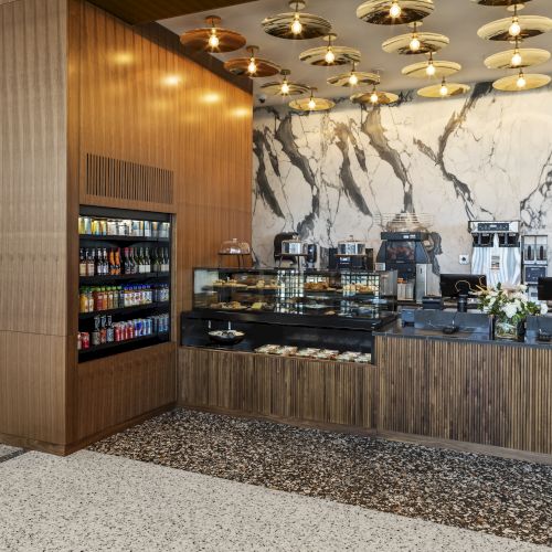 The image shows a sleek cafe setup with a beverage fridge, pastry display, coffee machines, and elegant lighting.