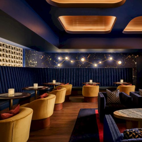 A stylish lounge with blue velvet walls, warm amber lighting, and plush yellow chairs around small round tables, perfect for cocktails.