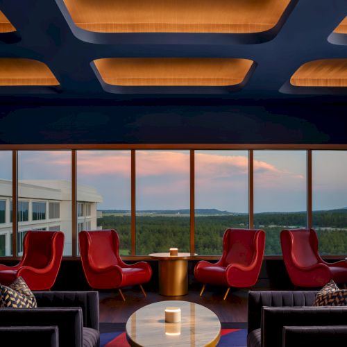 A stylish lounge with red armchairs, a glass coffee table, and a panoramic window overlooking a green landscape under a warm ceiling glow.