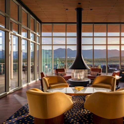 A modern lounge with warm yellow chairs around a central fire pit, large glass walls, and a sunset view over distant hills.