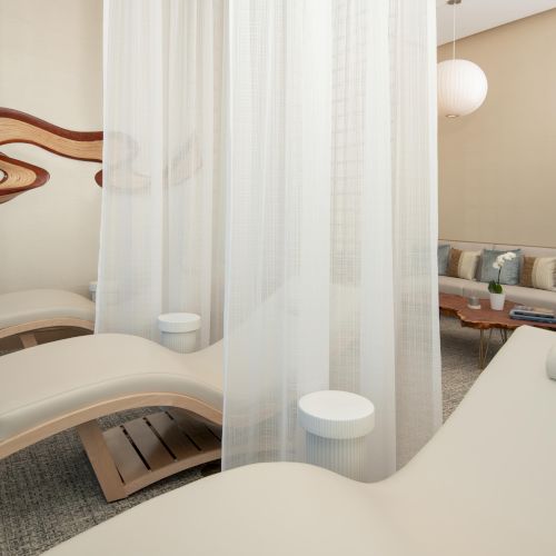 The image shows a serene spa-like room with lounge chairs, white curtains, and elegant decor including a wooden wall accent and soft lighting.