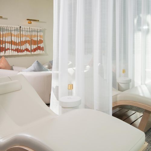 The image shows a bright, modern relaxation space with lounge chairs, soft curtains, and a decorated wall accent.