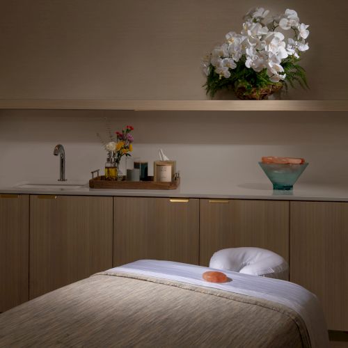 A spa room with a massage table, decorated with white orchids, a tray of candles, a sink, and a bowl of stones on the counter.