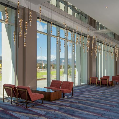 A modern lobby with tall windows, stylish lighting, red chairs, and a geometric carpet, overlooking a scenic outdoor view.