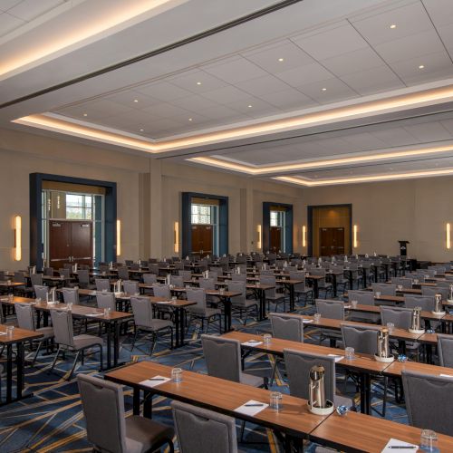 A conference room setup with rows of chairs and tables, equipped with notebooks, glasses, and water pitchers, under modern lighting.