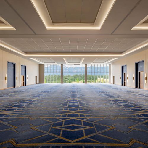 A spacious, well-lit hotel conference hall with patterned blue carpet, modern lighting, and large glass doors opening to greenery outside. (ends with period)