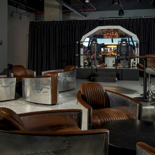 A lounge area with leather and metal chairs faces a room featuring a flight simulator cockpit setup.