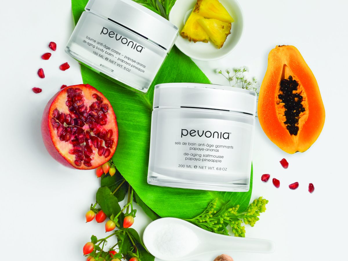 Pevonia anti-age body balm and bath salts in jars with papaya, pineapple, pomegranate, and green botanicals on a white background.